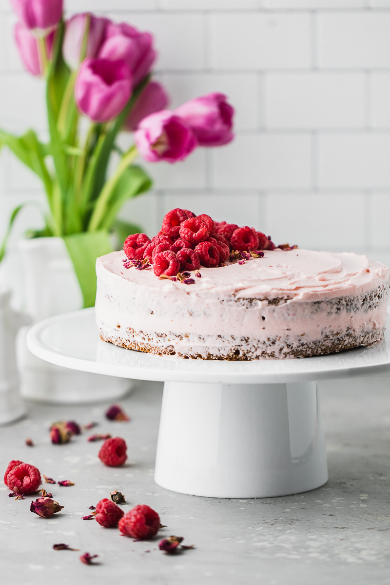 Raspberry frosted carrot cake on white cake stand and flowered in the background