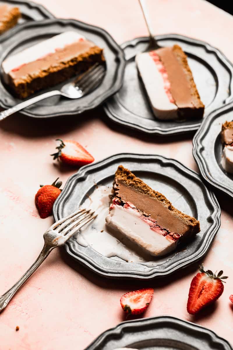 Slices of dairy free ice cream cake with layers of chocolate and strawberry on plates and a pink surface.