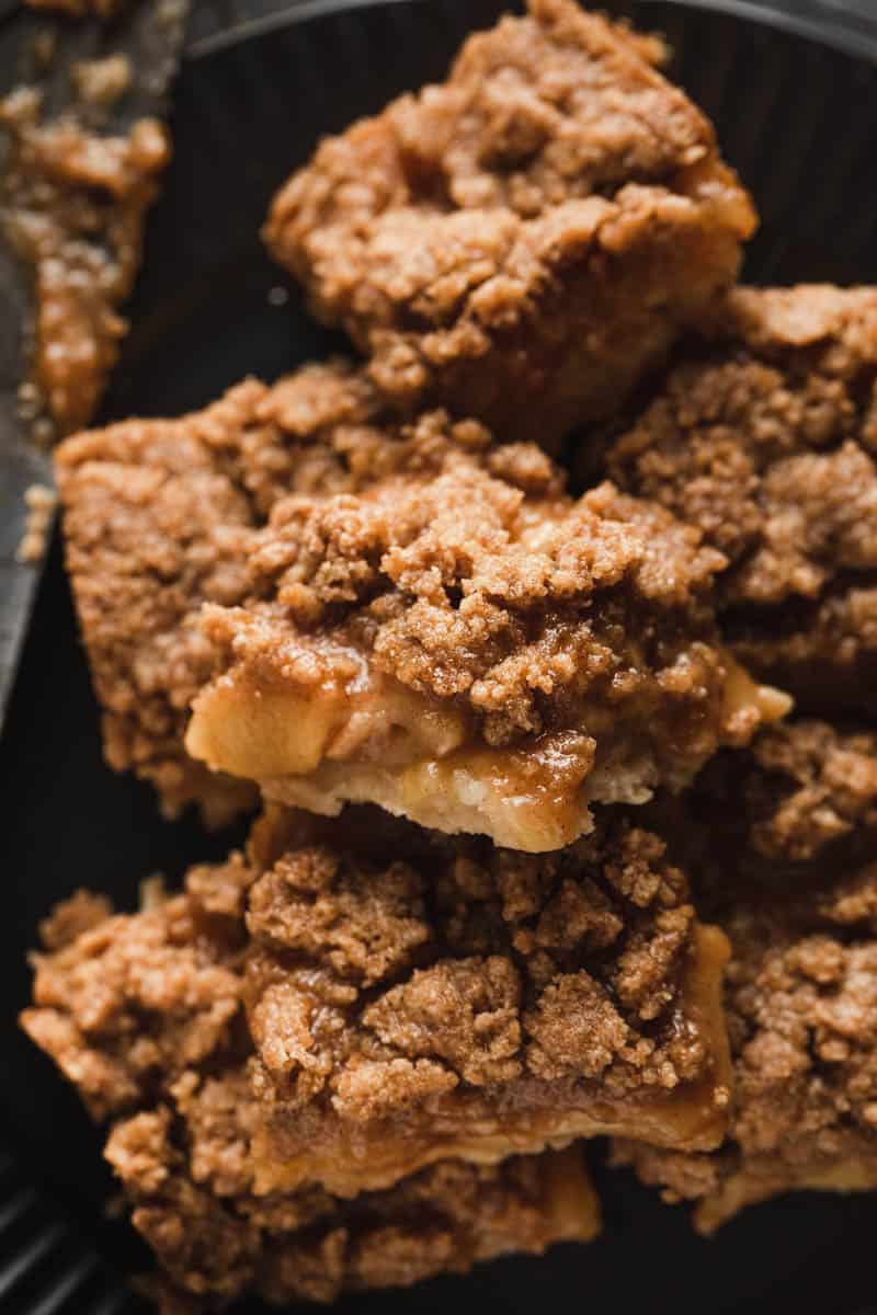 apple crumble pie bars stacked on a black plate with warm apple pie filling spilling out. 